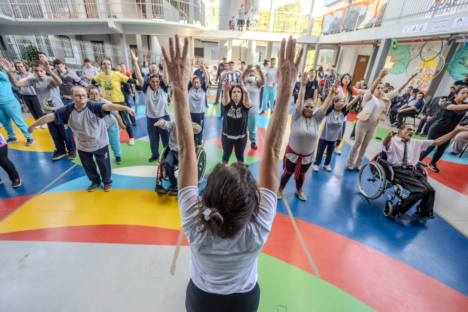 Foto horizontal de uma aula da Lápis de Seda Cia de Dança. A imagem mostra uma mulher de costas para foto com os braços erguidos para cima. À frente dela estão dezenas de pessoas de diferentes idades e características físicas. Duas pessoas estão em cadeiras de rodas. Todos estão dentro de um ginásio com o chão colorido.