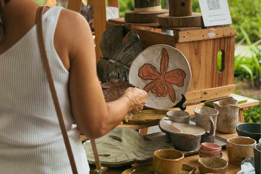 Foto horizontal mostra pratos e xícaras de cerâmica coloridos expostos em uma mesa de madeira no Festival Meraki, na Praça Cidade das Águas. No lado esquerdo da foto, aparece parcialmente o tronco de uma mulher, de costas para a foto e de frente para os materiais expostos.  