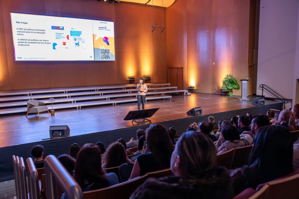Foto horizontal mostra um mulher de meia-idade, de cabelos pretos falando ao microfone em um grande palco de madeira. Atrás dela, um grande telão com uma apresentação. À frente dela, e de costas para foto, centenas de pessoas sentadas em cadeiras estofadas em uma plateia, disposta em estilo arena, com vários degraus. 