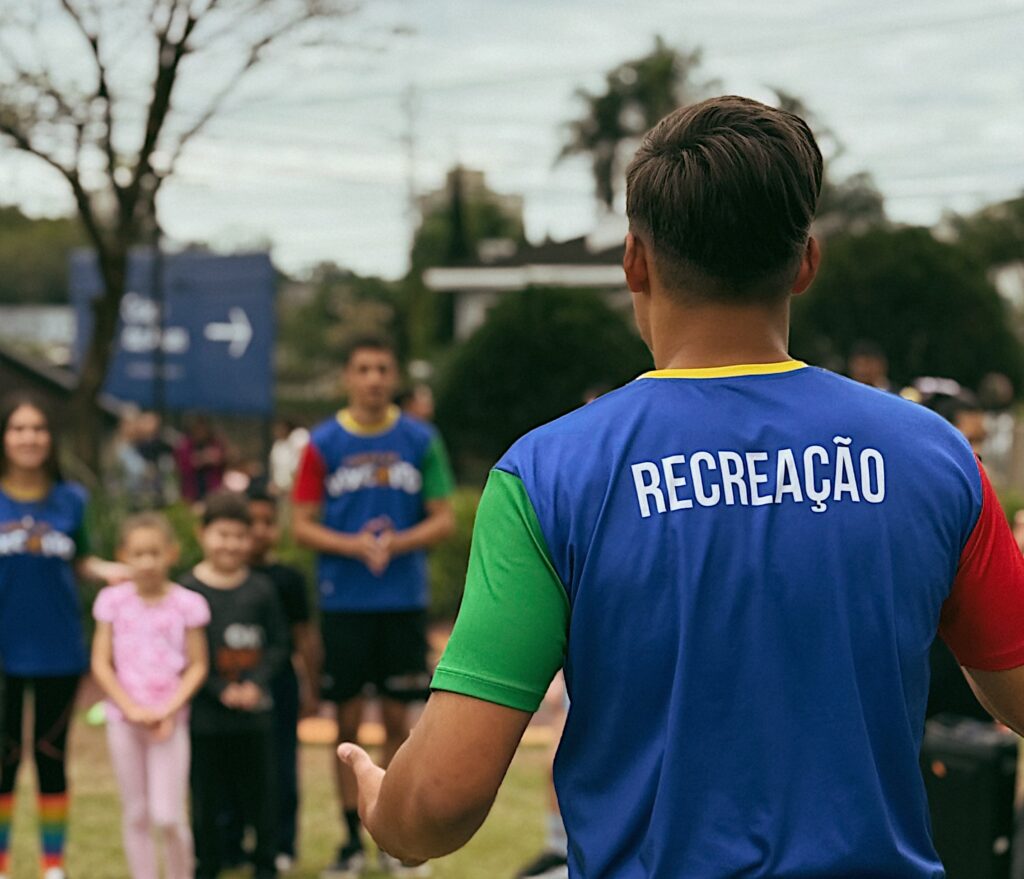 Foto horizontal mostra um homem de costas. Ele usa uma camiseta azul com manga verde. Nas costas está escrito "Recreação". Ao fundo, crianças e adultos desfocados. Eles estão na Praça Cidade das Águas, em Joinville. 