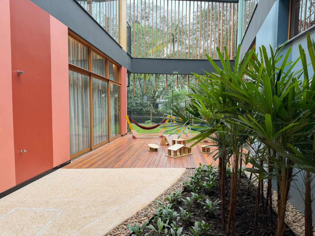 Foto horizontal mostra ambiente coberto entre duas salas da unidade infantil da Coree International School, com arbustos e espaço ao ar livre no fundo. 