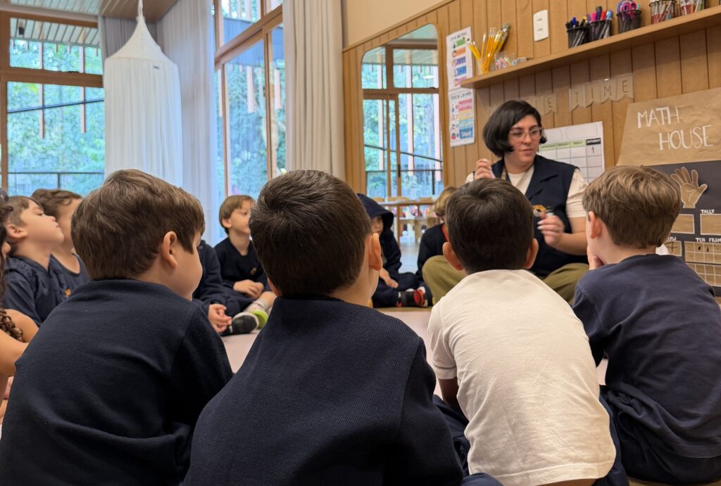 Foto horizontal mostra nove crianças sentadas em roda à frente de uma professora em uma das salas de educação infantil da Coree International School 