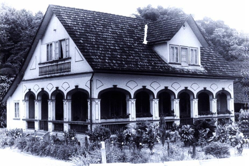 Foto horizontal, em preto e branco, mostra a fachada da Casa Krüger, um casarão com uma grande varanda, com dez arcos ornamentados e balaustres. A casa tem um sótão e telhado estilo duas águas. Na frente da casa há canteiros com plantas ornamentais e ao fundo aparecem algumas árvores.