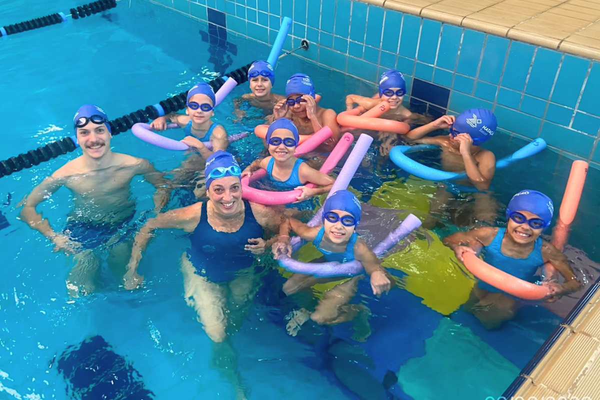 Foto na horizontal mostra oito crianças de idades entre 7 e 10 anos dentro de uma grande piscina, no complexo esportivo da Coree International School. Elas usam maiô e sunga na cor azul clara, toucas e óculos de natação em azul escuro. Todas estão envolvidas com boias compridas. Elas estão acompanhadas por dois professores de natação, um homem jovem e uma mulher de meia-idade, ambos de pele clara. Todos olham para a foto e sorriem. 
