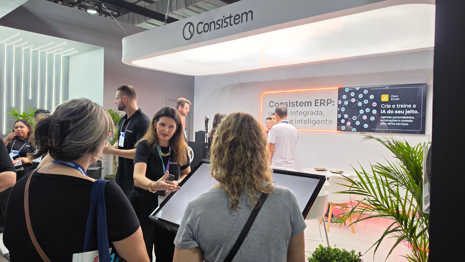 Foto na horizontal mostra o estande da Consistem na Febratex. O espaço tem paredes brancas, mesas e cadeiras claras. Em uma das paredes há um painel com alguns informações, que aparecem parcialmente: "Consistem ERP. Crie e treine a IA do seu jeito". À frente do estande duas mulheres, de costas para foto, conversam com uma mulher de meia-idade e uma blusa preta da Consistem, que gesticula em frente a um totem. 