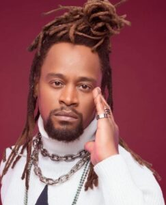 Foto de Toni Garrido. Ele é um homem preto, de meia-idade, rosto alongado, olhos castanhos, lábios grossos, barba e bigode curtos. Seu cabelo tem dreads longos. Ele usa uma blusa de gola alta branca e várias correntes de elo prateadas. O fundo é cor de vinho. 