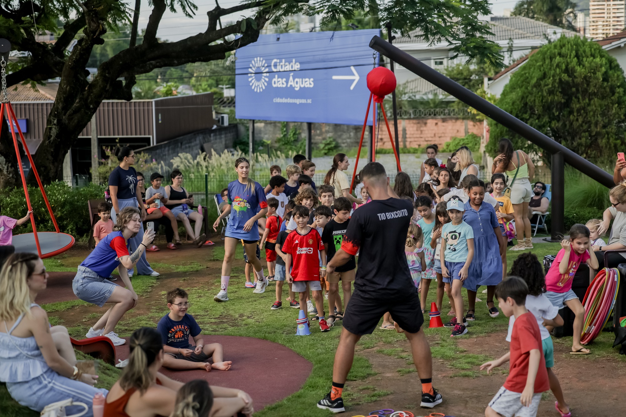Foto na horizontal mostra dezenas de crianças, de diferentes idades e característias físicas no gramado da Praça Cidade das Águas. Algumas estão em pé e outras sentadas. Aparecem alguns adultos sentados também. À frente de um grupo de crianças há um homem adulto, de pele parda e de costas para a foto. Ele usa uma camiseta preta escrito Tio Bixcoito nas costas. Ao fundo, aparecem algumas árvores e um grande painel azul escrito Cidade das Águas. 