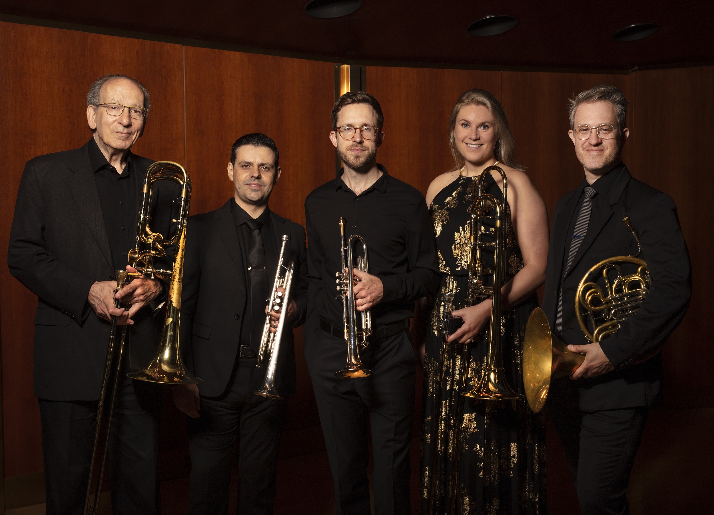 Foto na horizontal de cinco pessoas: quatro homens e uma mulher, integrantes do American Brass Quintet. Todos eles têm pele clara. Eles estão em pé, com roupas pretas, seguram seus instrumentos musicais, olham para foto e sorriem discretamente. Ao fundo, uma parede de madeira.