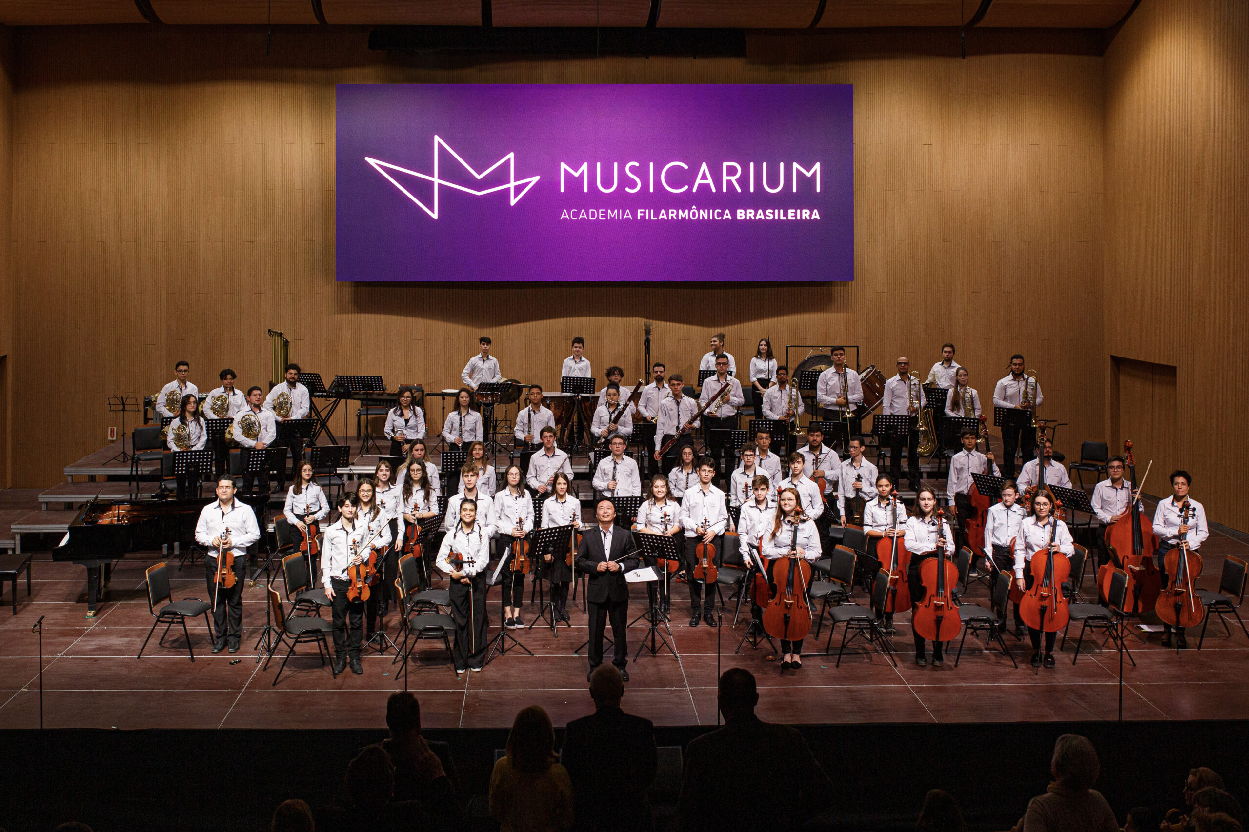 Foto horizontal mostra cerca de 50 músicos jovens em um grande palco. Eles estão em pé segurando seus instrumentos. À frente deles, o maestro Sergio Ogawa. Ele é um homem de meia-idade, média estatura, magro e com traços fisionômicos orientais. No fundo do palco, uma placa roxa com a logomarca do Musicarium.