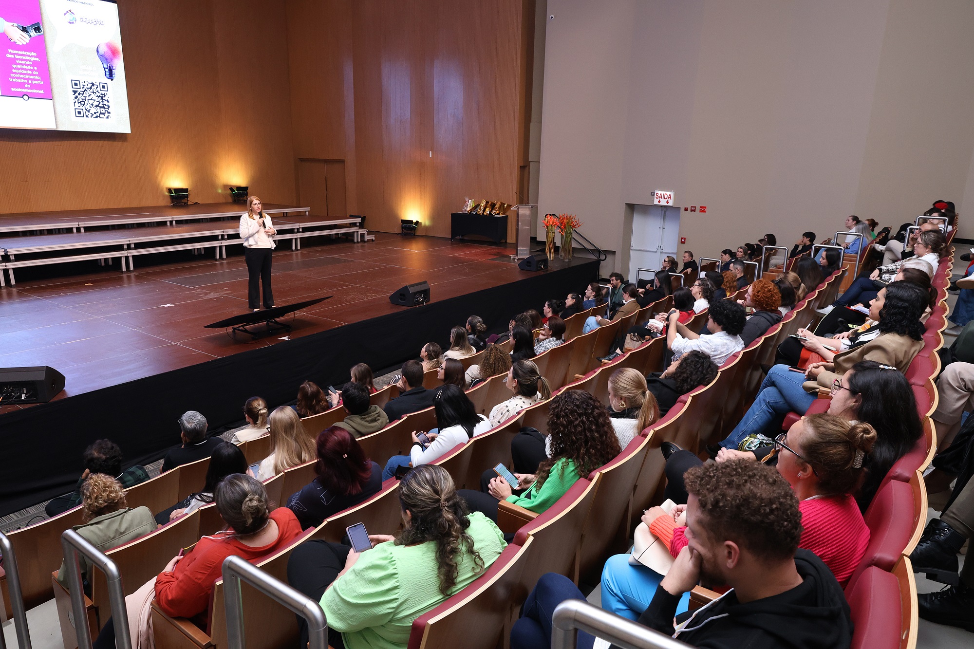 Foto horizontal mostra o grande auditório da Coree Internacional School. Sobre o palco está uma mulher adulta falando ao microfone, durante o Seminário de Educação Inovadora. De frente para ela e de costas para foto uma plateia lotada de pessoas.