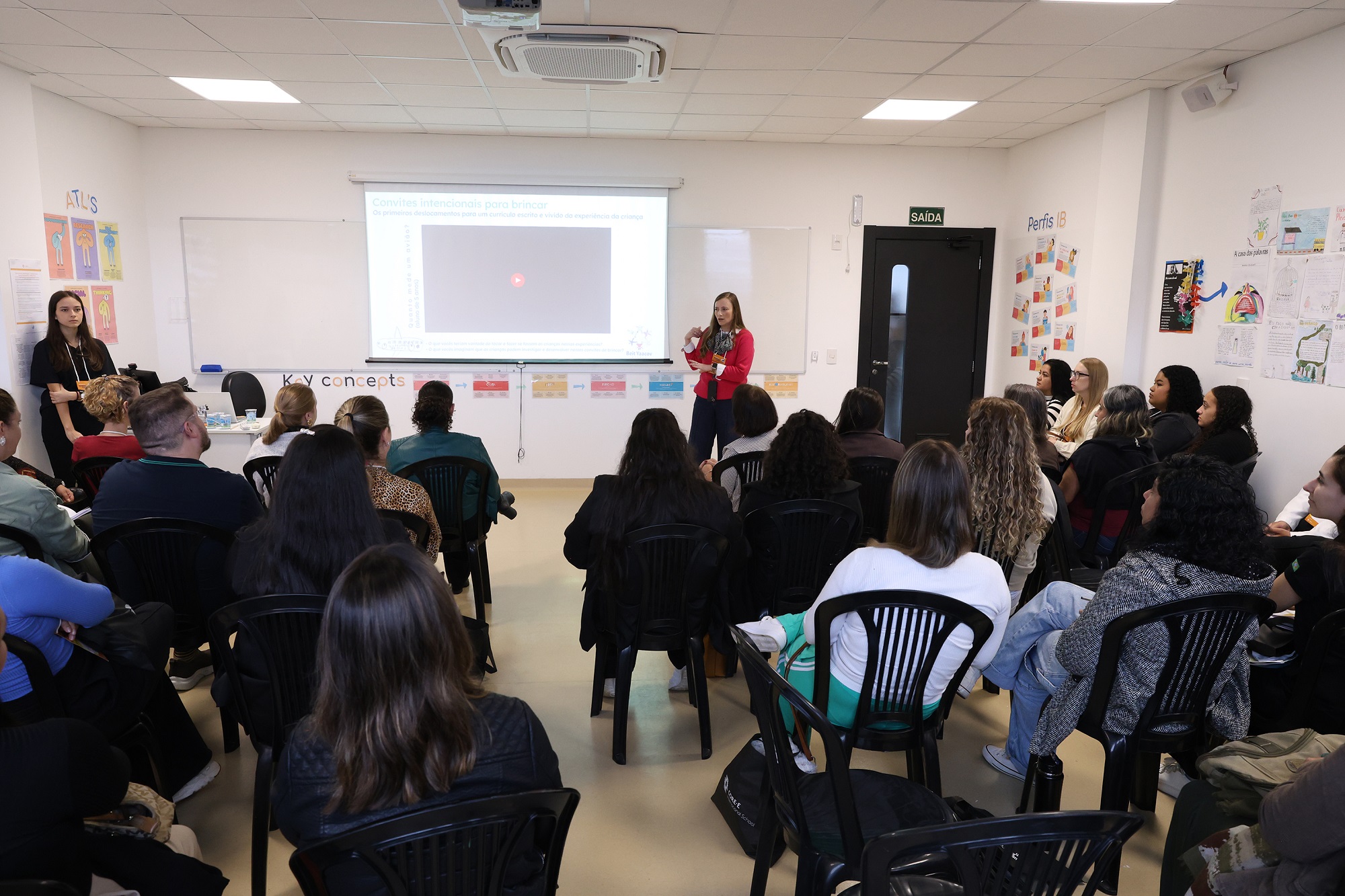Foto horizontal mostra uma sala de aula com cadeiras pretas e uma mulher em pé gesticulando ao lado de uma projeção. Dezenas de pessoas assistem, de costas para a foto.