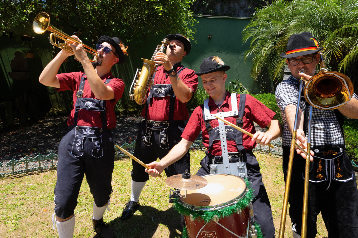 Foto horizontal mostra quatro homens jovens vestidos com roupas folclóricas germânicas - camisa social vermelha, supensório e calça corsário pretos. Cada um faz uma pose tocando um instrumento musical. Eles estão em um ambiente externo, sobre um gramado. Ao fundo, muro verde e árvores.