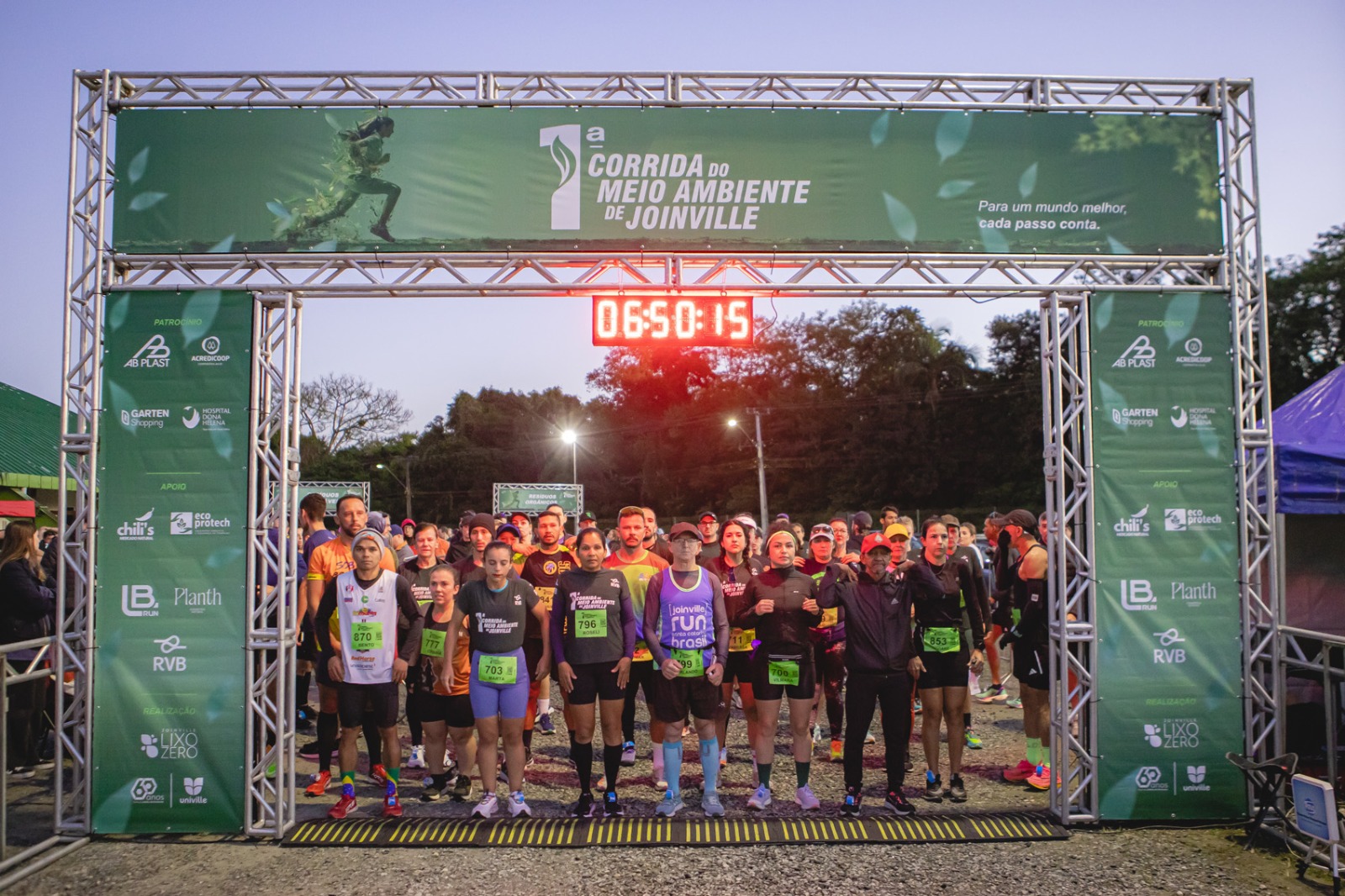 Foto horizontal mostra dezenas de pessoas embaixo de uma grande estrutura metálica com um lona verde da 1ª Corrida do Meio, que é a largada da corrida.