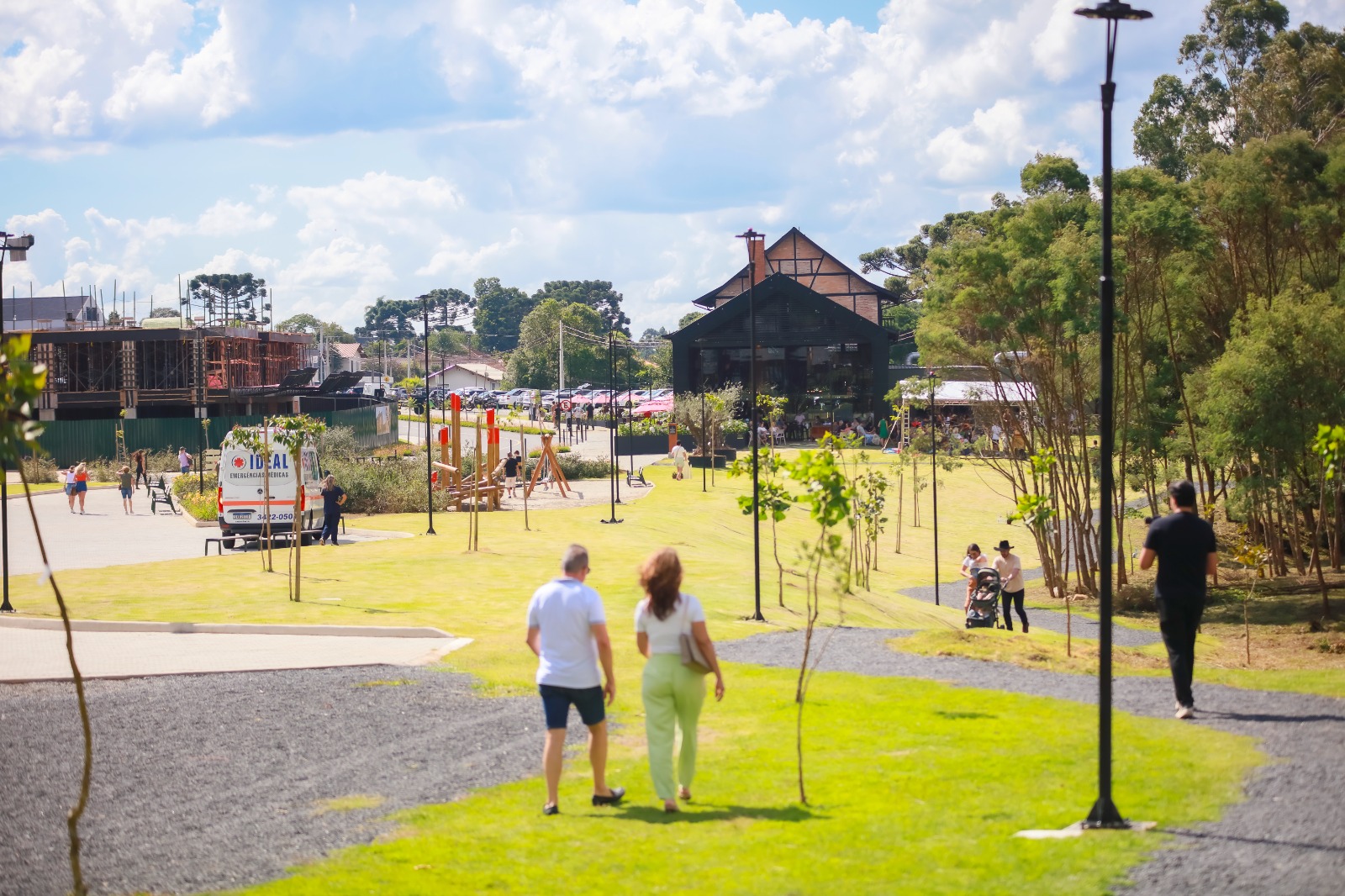 Foto horizontal mostra o Parque Paraíso das Araucárias - uma ampla área gramada com algumas árvores e várias pessoas caminhando. Ao fundo, aparece a estrutura do Casarão, um complexo gastronômico instalado em um edifício horizontal enxaimel.