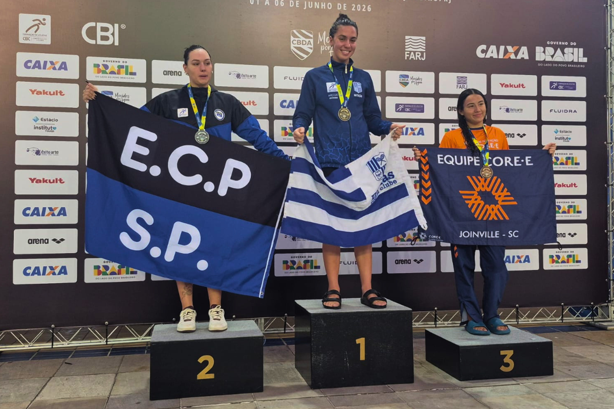 Foto horizontal mostra três mulheres jovens em cima de um pódio. Elas seguram as bandeiras de suas instituições. Atrás delas, um grande painel com o nome do evento e logomarcas. À direita na foto, a atleta Maria Laura ocupa o terceiro lugar do pódio. Ela é uma mulher jovem, de pele parda, cabelos longos e pretos. Ela segura uma bandeira azul com a logomarca laranja da Coree International School e os escritos: Equipe Core-e - Joinville - SC.