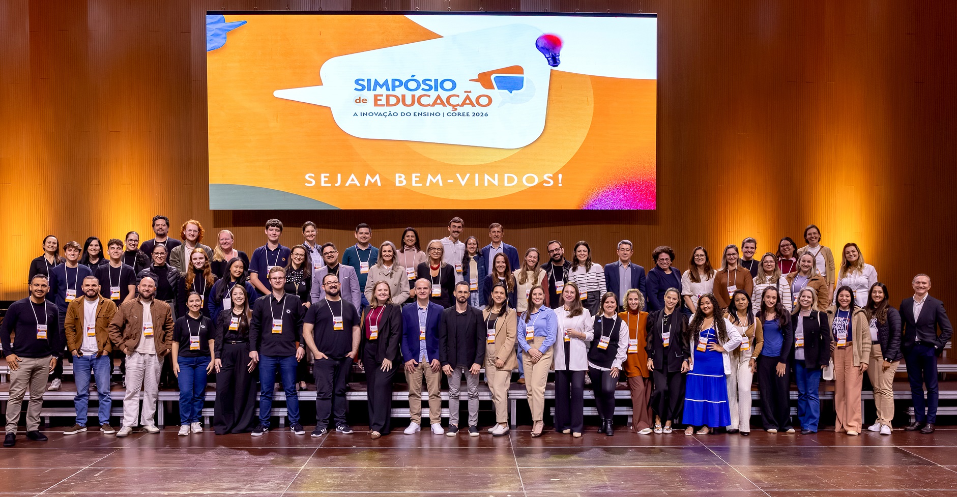 Foto horizontal mostra dezenas de pessoas em cima de um grande palco de madeira posando para a foto. Atrás delas, um grande telão onde está escrito Simpósio de Educação