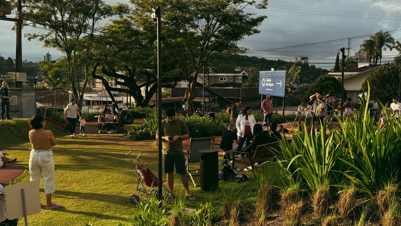 Foto horizontal mostra um amplo gramado, com arbustos, árvores e algumas pessoas em pé. Ao fundo, uma placa azul onde está escrito Cidade das Águas