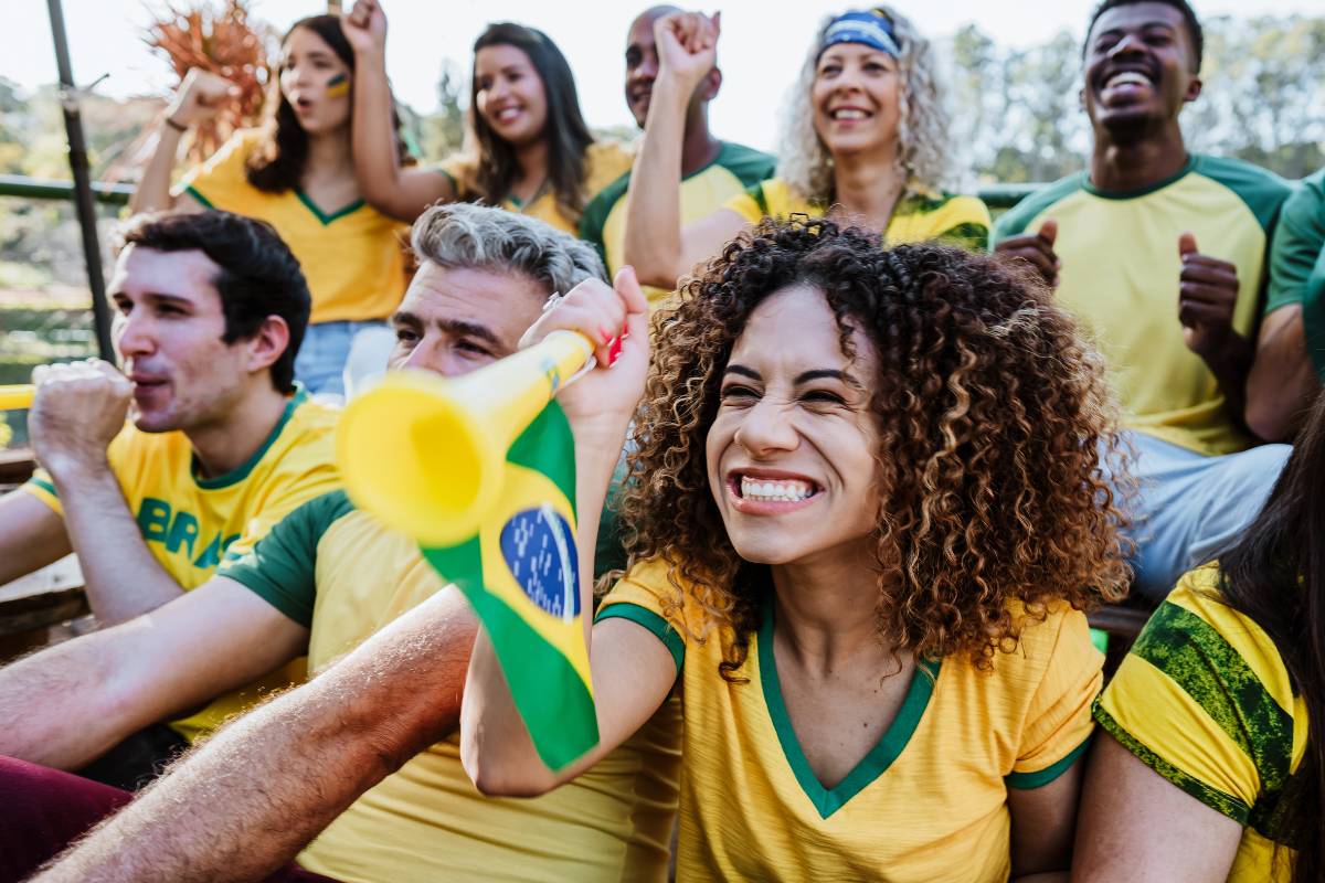 Foto horizontal mostra oito pessoas de diferentes idades e características físicas. Elas usam camisetas amarelas com detalhes em verde. Elas olham para frente e sorriem. Algumas estão com o punho cerrado. Uma mulher segura uma corneta com a bandeira do Brasil. Eles estão em um ambiente externo. O fundo é desfocado. A imagem ilustra o fato de a Praça Cidade das Águas, em Joinville, disponibilizar um telão para transmissão dos jogos do Brasil durante a Copa do Mundo.