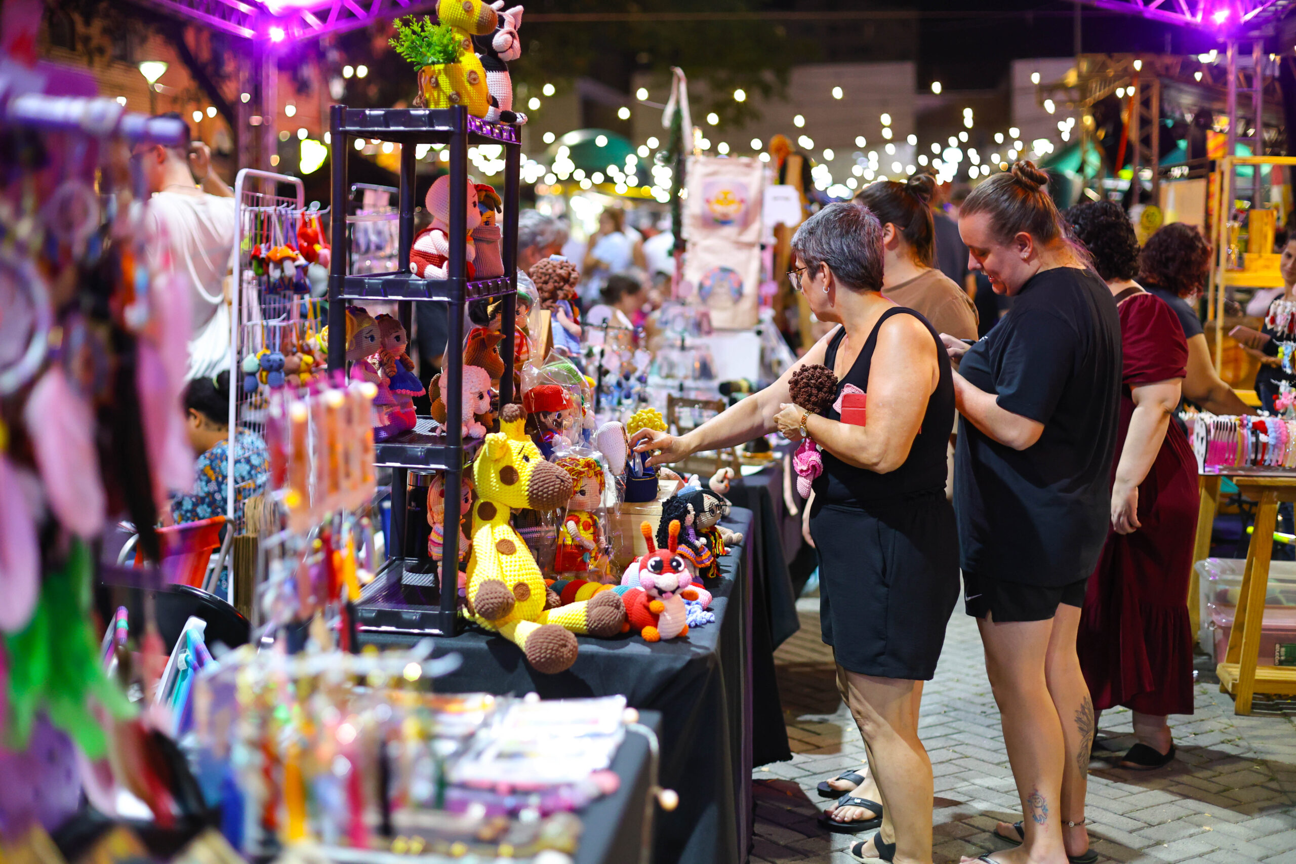 Foto horizontal de uma das edições do Viva Noite! Mercado Criativo. A imagem mostra várias barracas, montadas na rua, com produtos à venda, como bonecos de crochê, itens de papelaria e acessórios. Algumas pessoas estão paradas em frente a uma das barracas e observam os produtos. Está de noite e há um varal de luzes ao fundo.