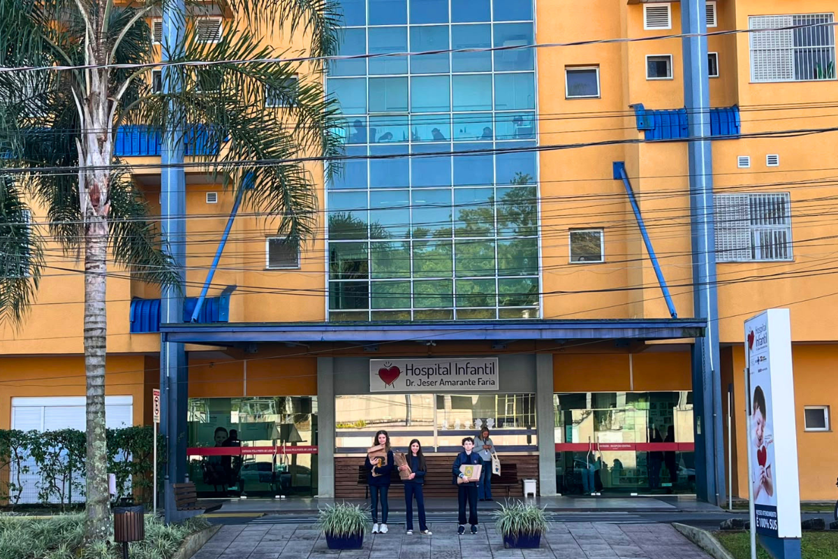Foto horizontal mostra a fachada do Hospital Infantil de Joinville - um prédio grande e alto, com paredes amarelas e vidros azulados. Em frente à entrada do hospital há três estudantes da Coree International School, de Joinville: duas meninas e um menino, ambos de pele clara e com idade em torno de 12 anos. Eles seguram envelopes pardos e olham para foto.