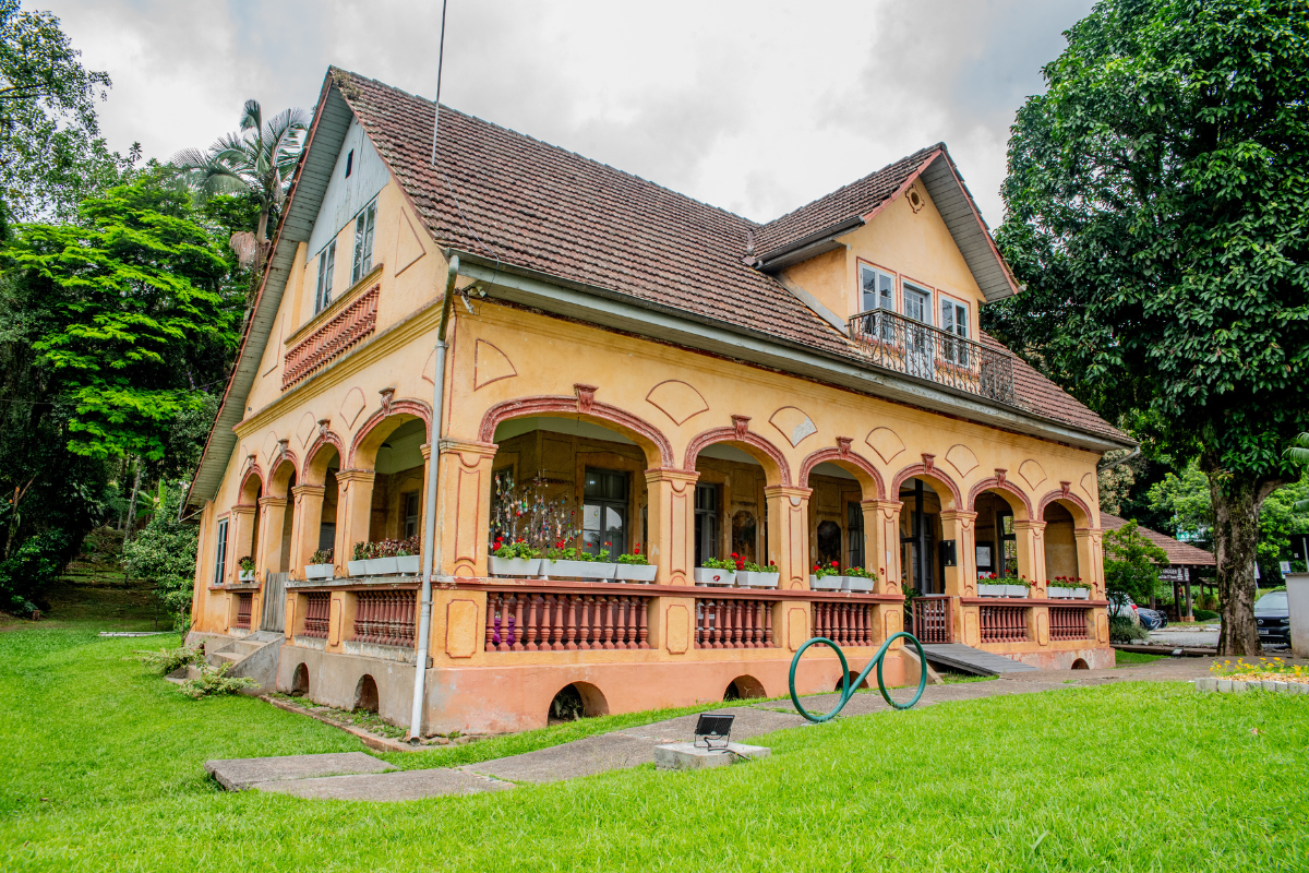 Foto horizontal mostra a fachada da Casa Krüger, um casarão com uma grande varanda, com dez arcos ornamentados, paredes na cor de amarelo envelhecido e balaustres marrons. A casa tem um sótão e telhado estilo duas águas. Na frente da casa há um grande gramado e ao redor aparecem parcialmente algumas árvores.