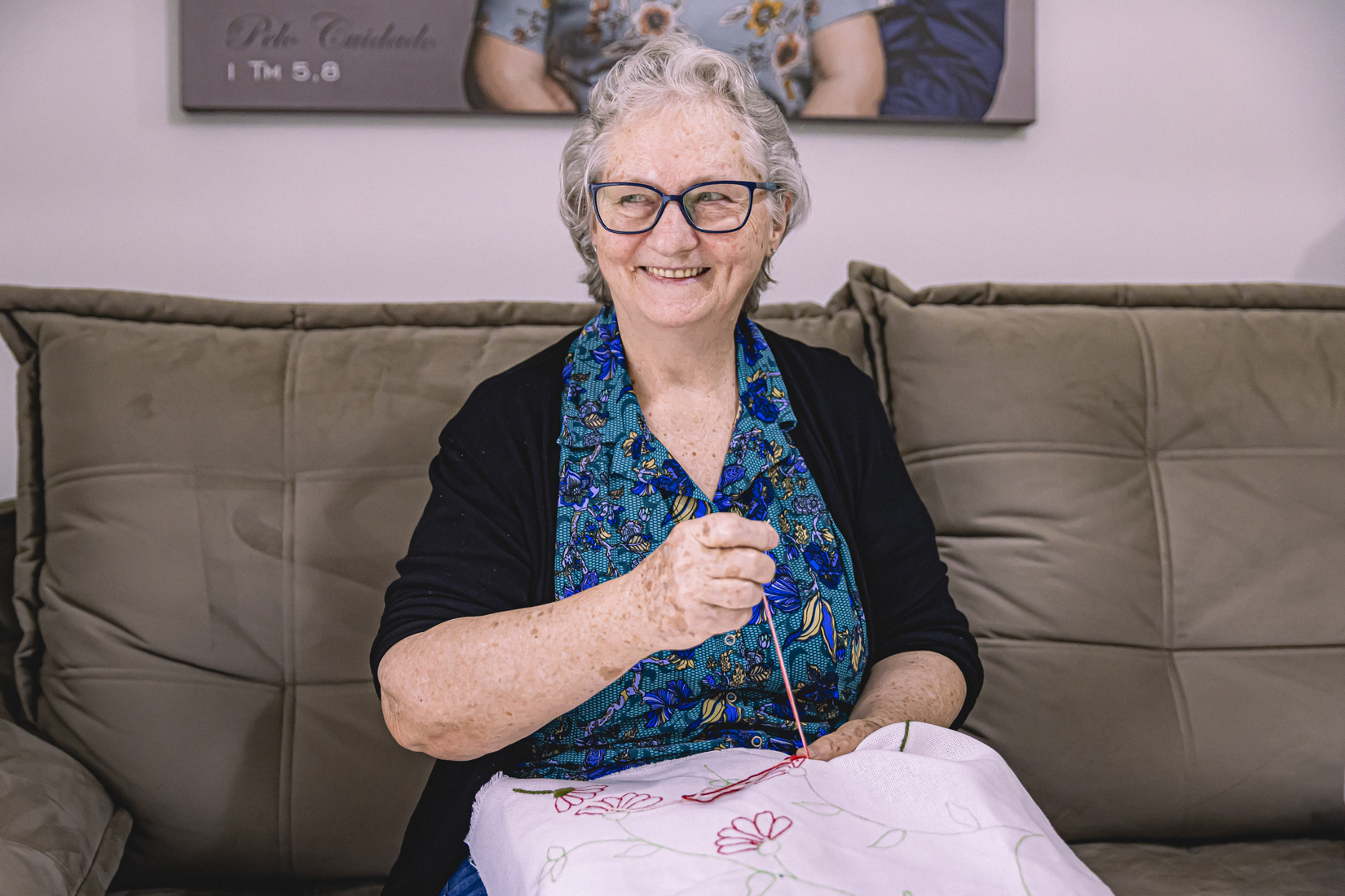 Foto horizontal mostra dona Lori Altmann. Ela é uma mulher idosa, de pele e olhos claros, cabelos brancos na altura do queixo. Ela usa um óculos retangular escuro e um vestido estampado com flores azuis. Está sentada em um sofá bege com um tecido no colo e uma linha esticada. Ela olha para o lado e sorri.