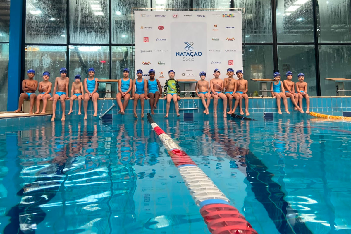 Foto na horizontal mostra em primeiro plano duas raias de uma grande piscina. Ao fundo, 16 crianças de idades entre 7 e 10 anos e diferentes características físicas estão sentadas na borda da piscina, com os pés em direção à água. Elas usam maiôs e sungas na cor azul claro, toucas em azul escuro e óculos de natação sobre as toucas. Atrás delas, há um grande painel branco com a logomarca do projeto Natação Social, a logomarca do Instituto Core e dos patrocinadores.