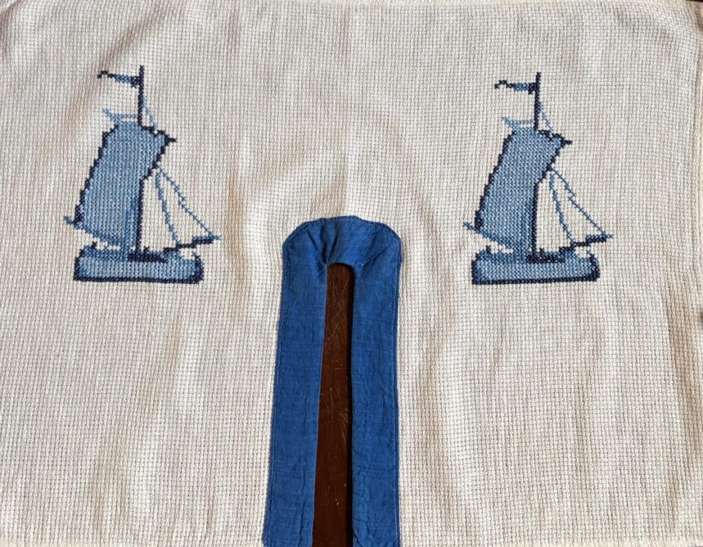 Foto horizontal mostra um tecido com branco com dois desenhos de um barco à vela, bordado em azul. No meio do tecido há uma fenda, contornada por um tecido azul.