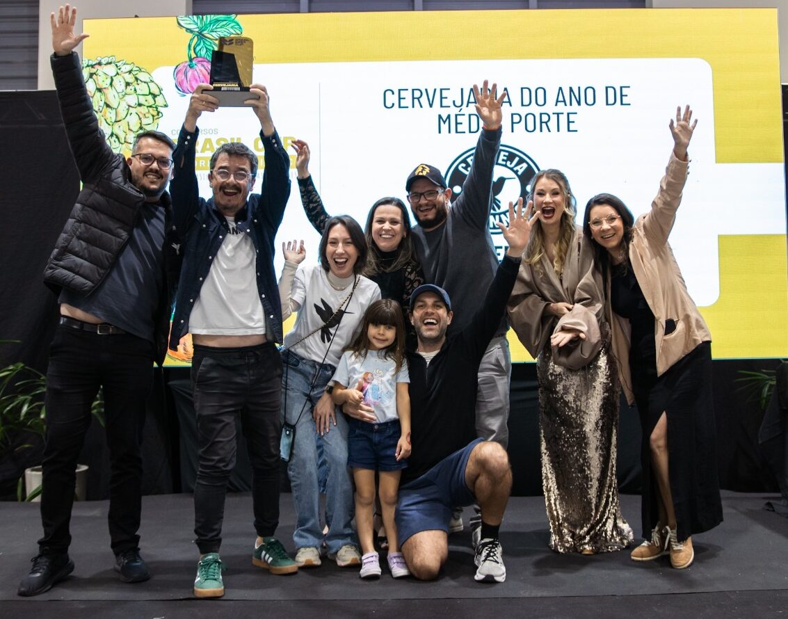 Foto horizontal mostra quatro homens e cinco mulheres, incluindo uma criança, de diferentes idades e características físicas. Eles estão em cima de um pequeo palco, sorriem e estão com as mãos para o alto. Atrás deles, um grande painel, onde aparece parcialmente o escrito: Cervejaria do ano de médio porte e abaixo a logomarca da Stannis, de Jaraguá do Sul.