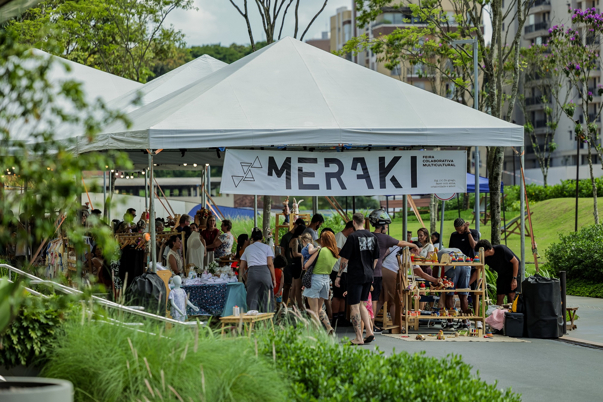 Foto horizontal mostra grandes tendas brancas instaladas na praça Cidade das Águas. Na frente da primeira tenda há uma faixa branca com a logomarca da Meraki e ao lado a informação: Feira Colaborativa Multicultural. Abaixo da tenda, dezenas de pessoas olham objetos expostos pelos expositores, entre eles brinquedos, roupas e acessórios. No canto esquerdo da foto, aparece parcialmente arbustos e folhagens da praça.