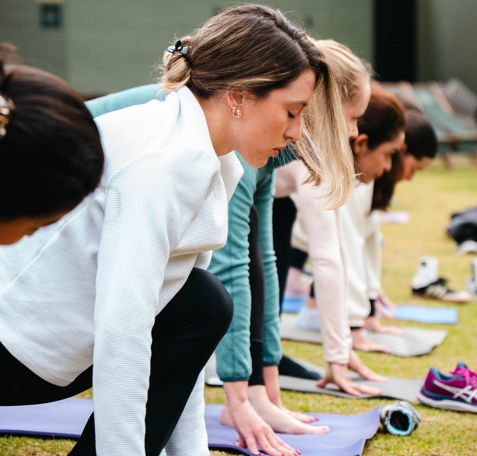 Foto horizontal mostra cinco mulheres jovens, de diferentes características físicas, uma lado da outra praticando yoga no gramado do Casarão Campo Alegre. Elas estão com a perna esquerda dobrada para frente e a perna direita esticada para trás e têm as duas mãos apoiadas em tapetinhos de yoga.