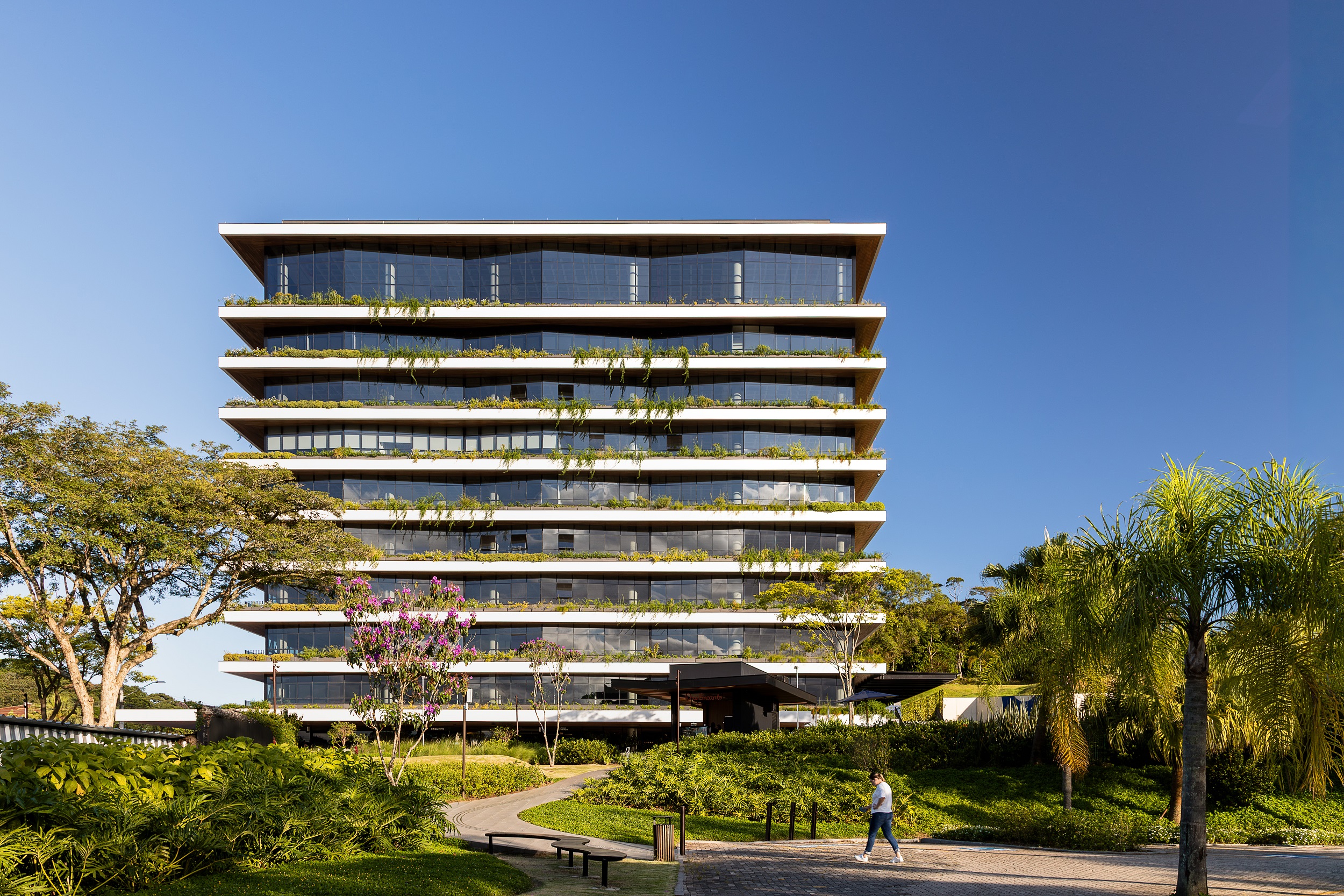 Foto horizontal mostra o Edifício Genève, de frente para a Praça Cidade das Águas. O prédio é largo, tem nove andares e vidros dispostos de forma diagonal. Em torno de cada andar há uma grande sacada com vegetação. À frente do prédio está a praça, com áreas gramadas, árvores e bancos. O céu está azul.